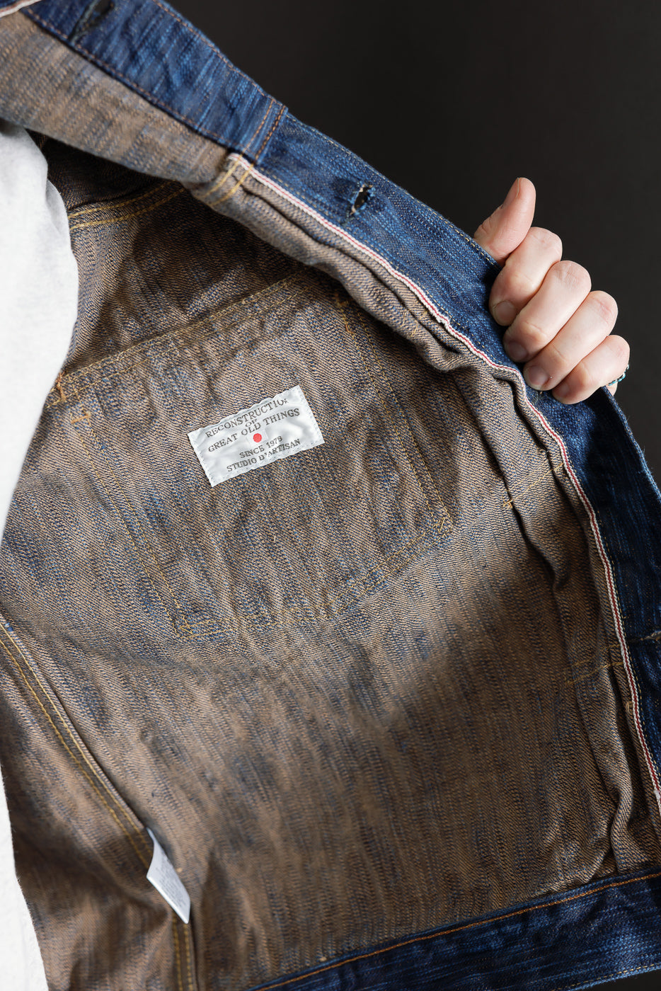Close-up of male model holding open natural indigo selvedge denim jacket, showing interior label and stitching.