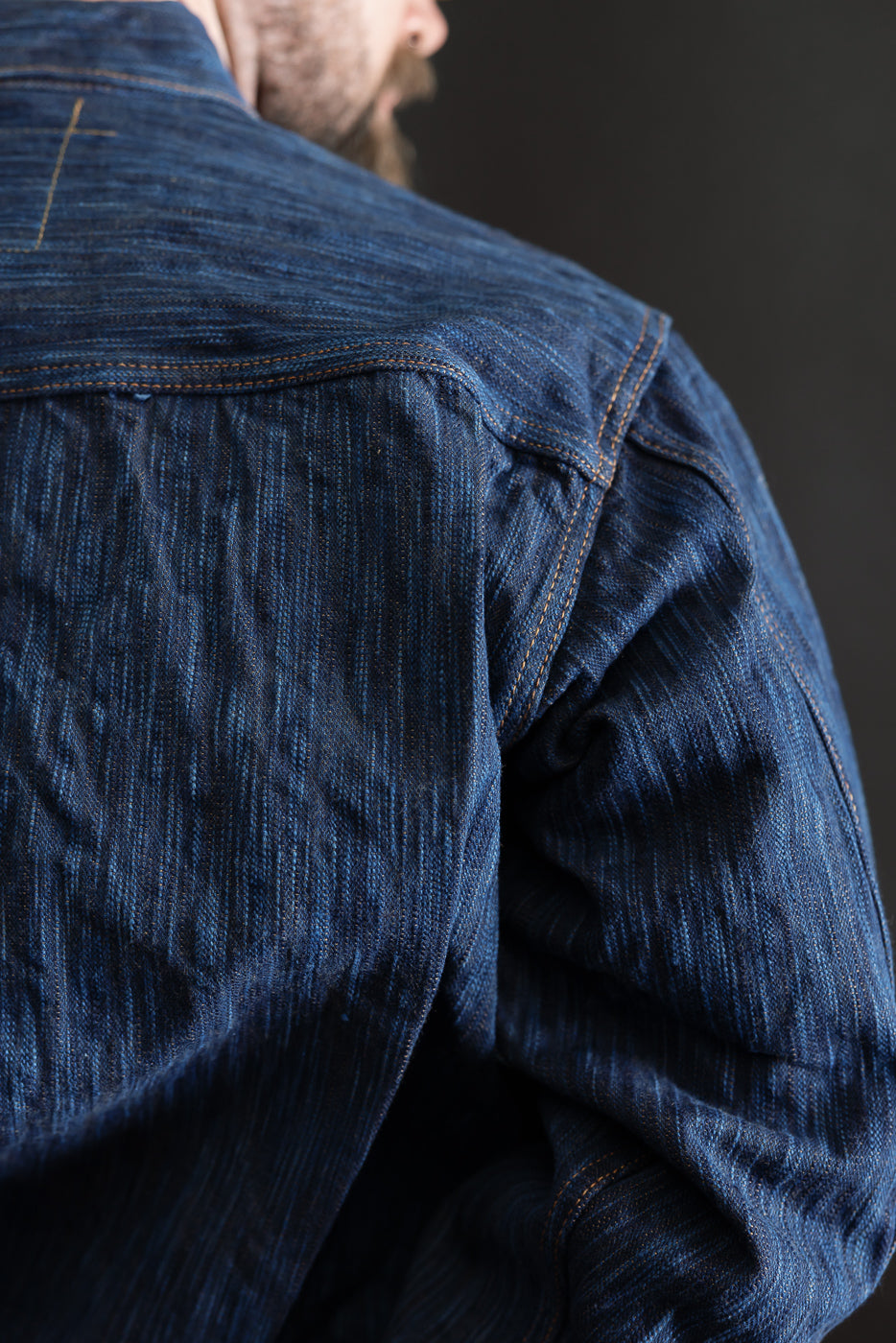 Close-up of male model's upper back and shoulder in natural indigo selvedge jacket, showing fabric texture and stitching details.
