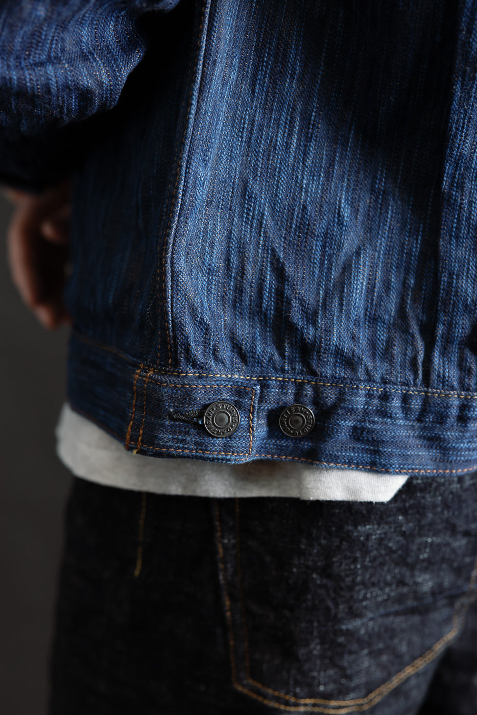 Close-up of male model's lower back in natural indigo selvedge denim jacket, showing side buttons and textured fabric. Model is 5'9