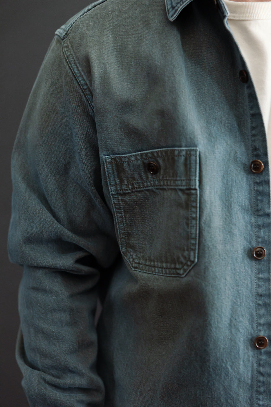 Close-up side view of a male model's teal denim shirt, showing the chest pocket and button placket.
