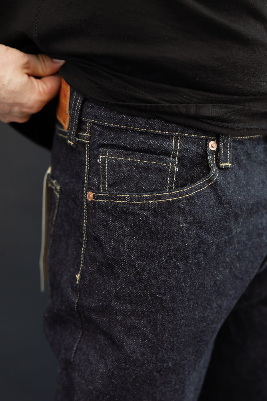 Close-up front view of dark selvedge denim jeans on male model, showing front pocket, copper rivets, and contrast stitching