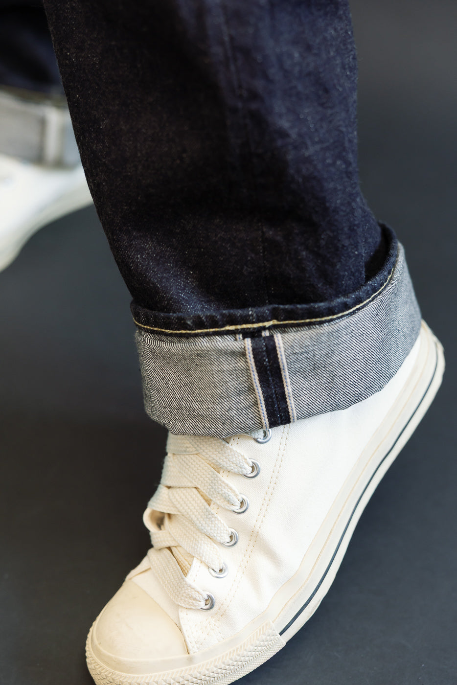 Close-up of male model’s cuffed selvedge denim hem with contrast stitching, paired with cream canvas sneakers