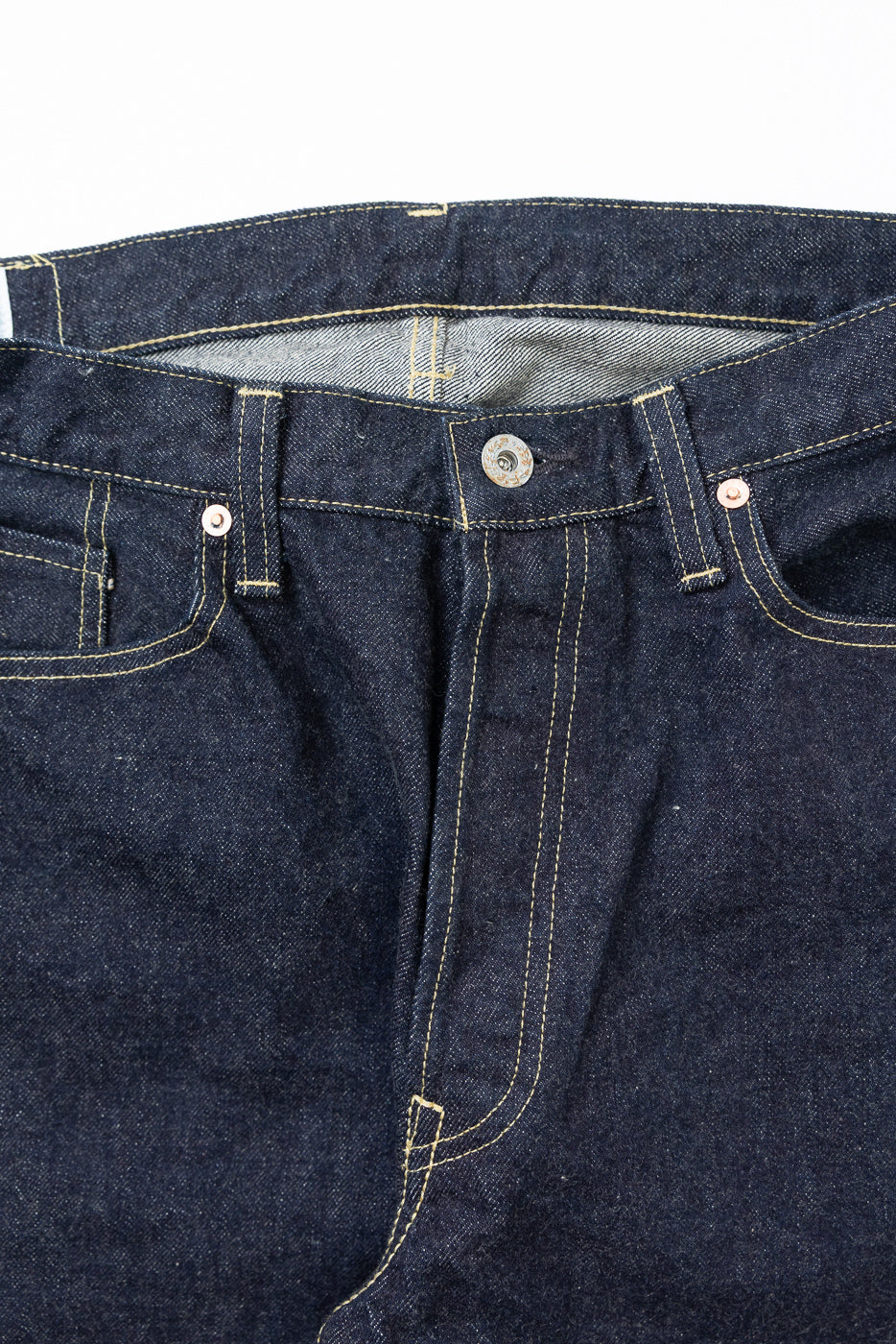 Close-up front view of dark selvedge denim jeans showing button fly, gold stitching, and fabric texture