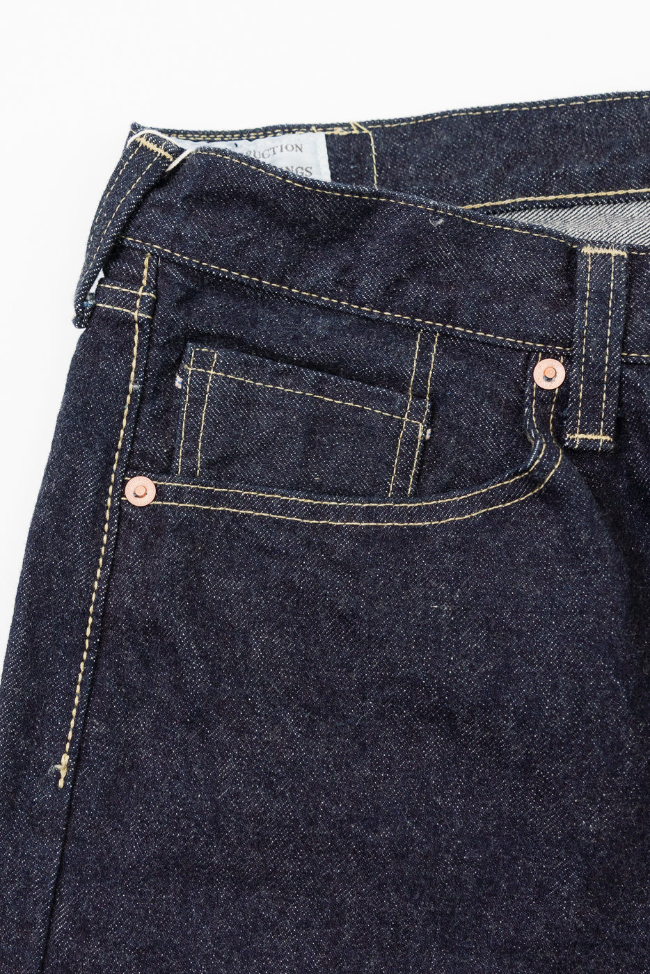 Close-up front view of dark selvedge denim jeans showing copper rivets, yellow stitching, and waistband tag detail