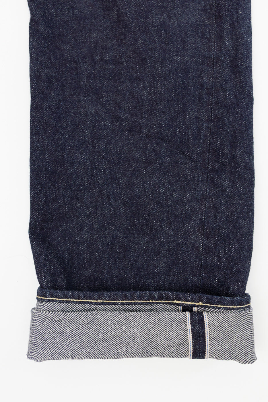 Close-up front view of dark selvedge denim jeans hem, showing raw edge, contrast stitching, and selvedge stripe detail