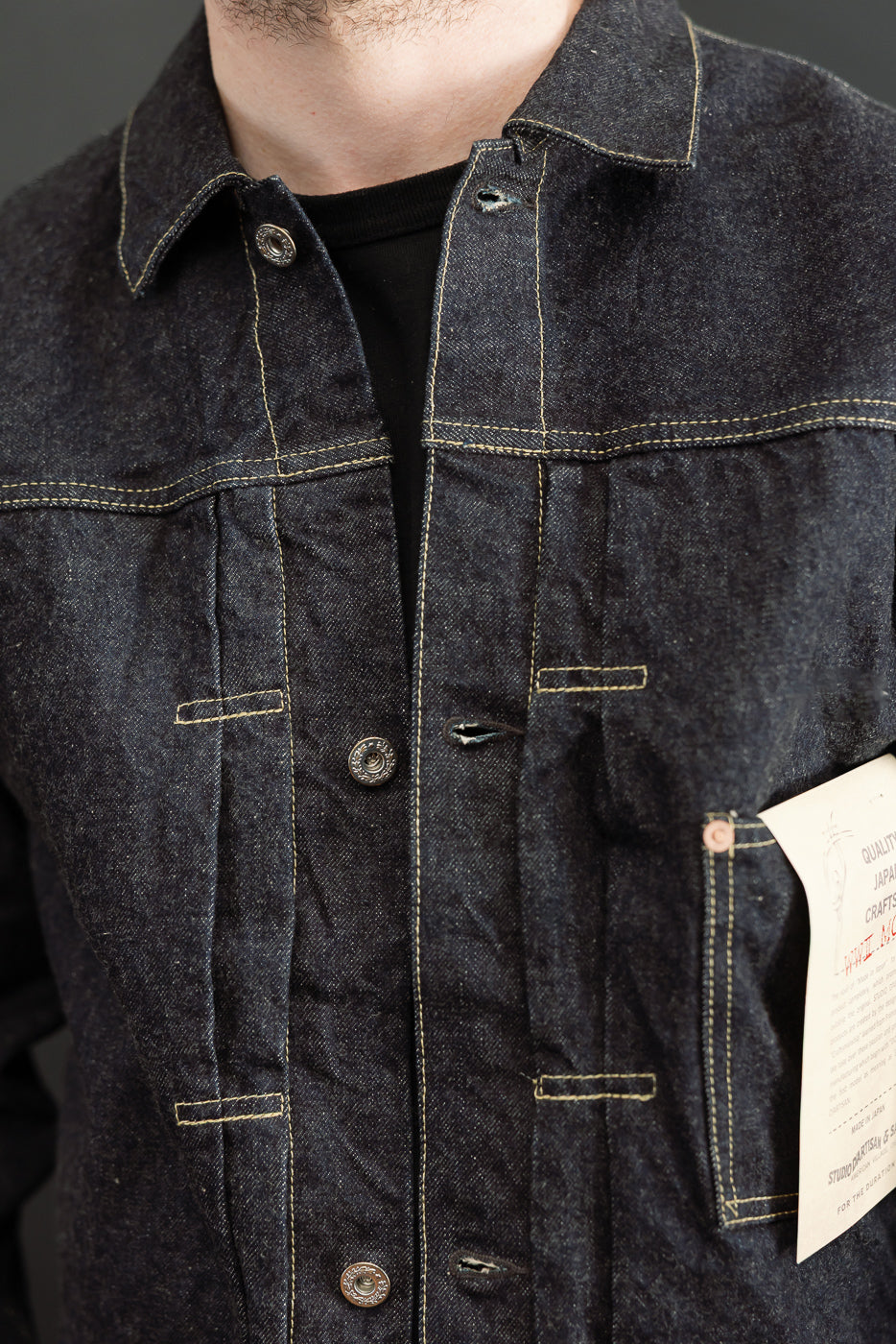 Close-up front view of male model wearing dark denim jacket with yellow stitching, buttons, and chest pockets.