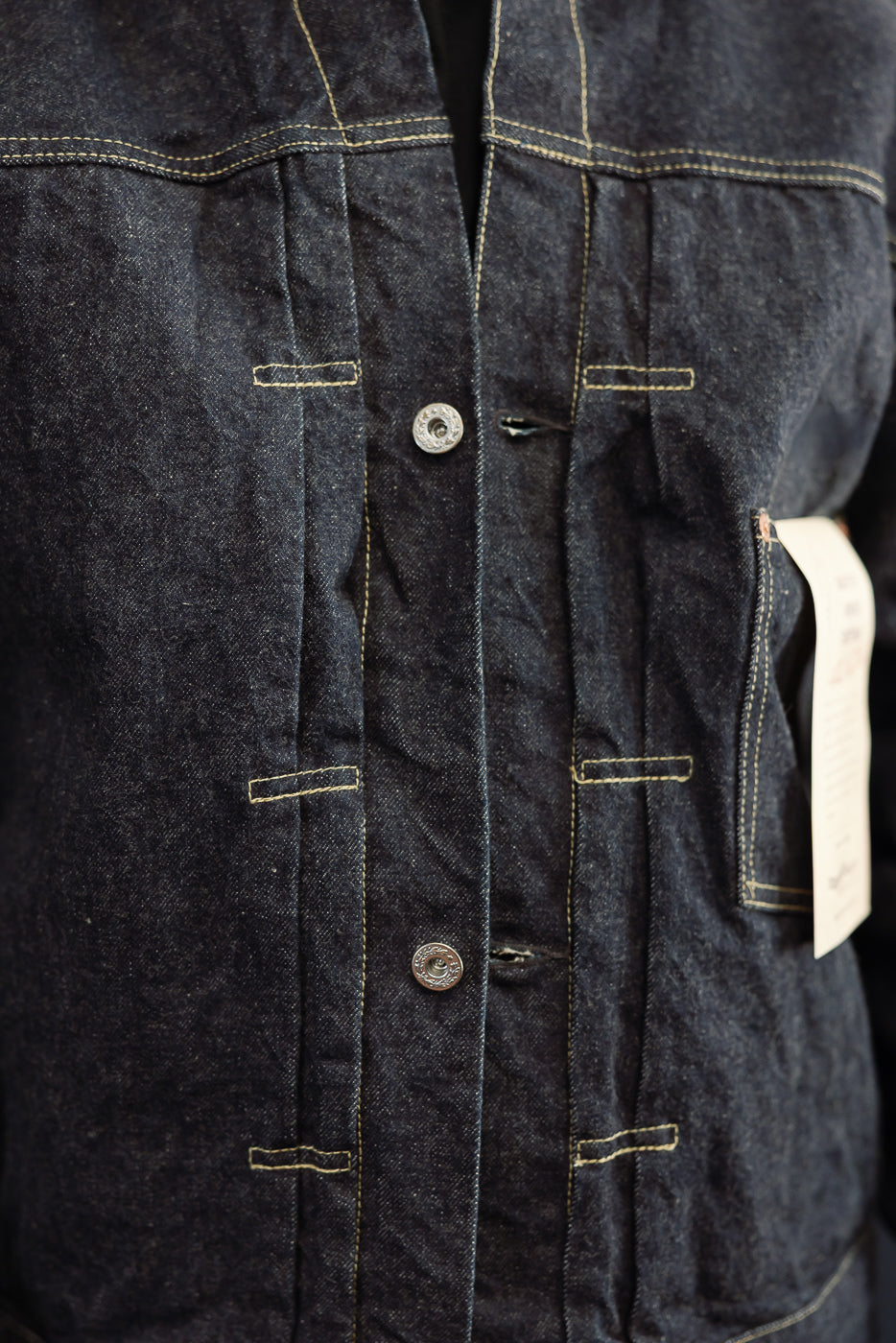 Close-up front of dark selvedge denim jacket, showing button placket, chest pockets, and contrast stitching.