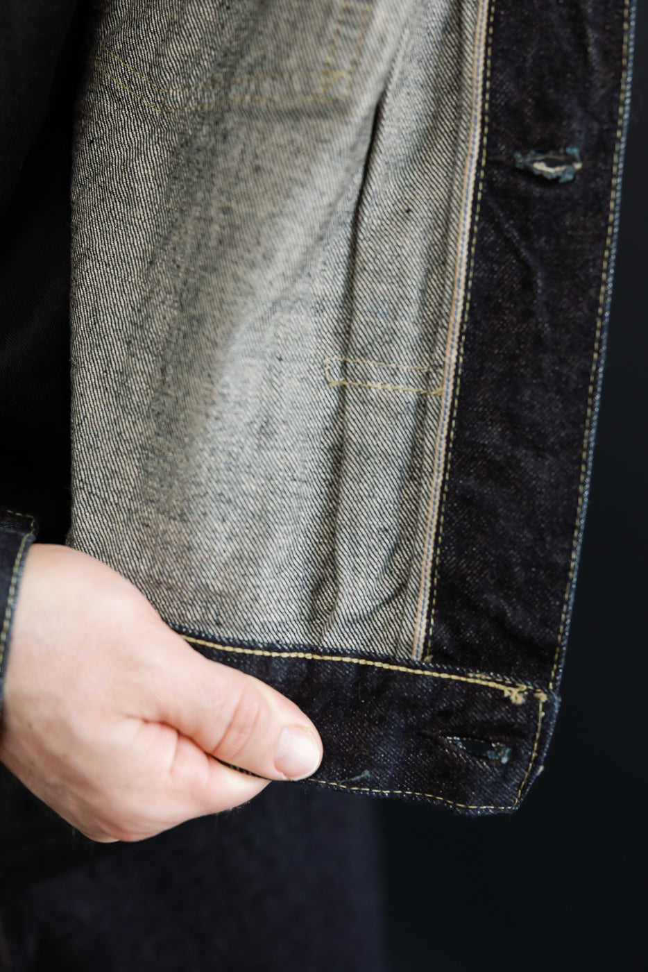 Close-up of selvedge denim jacket sleeve, front view, showing raw hem, contrast stitching, and fabric texture on male model