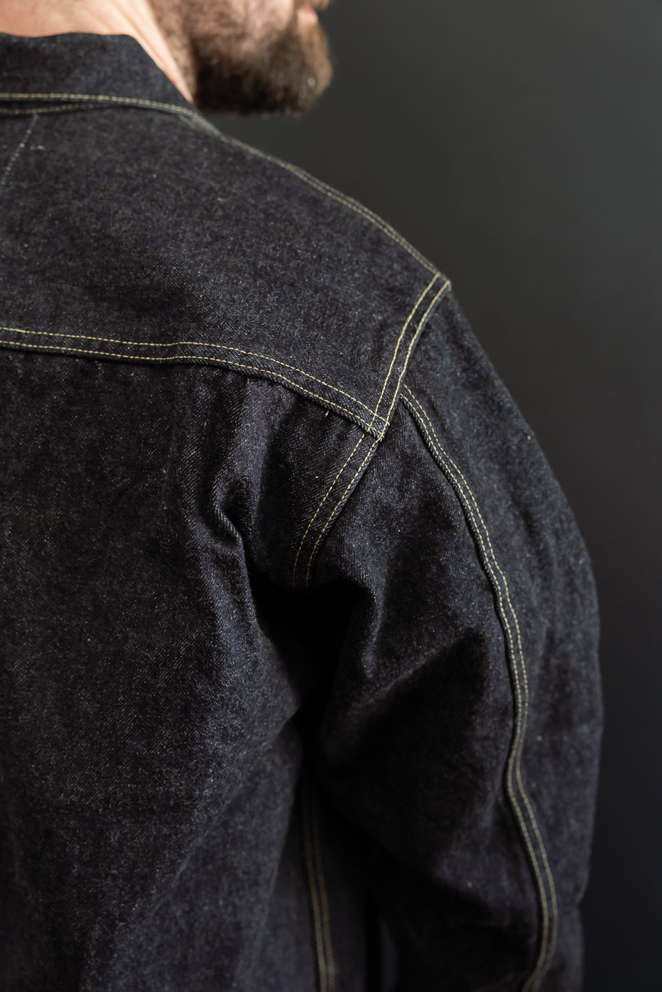 Close-up back view of male model in dark selvedge denim jacket, showing shoulder seam & contrast stitching