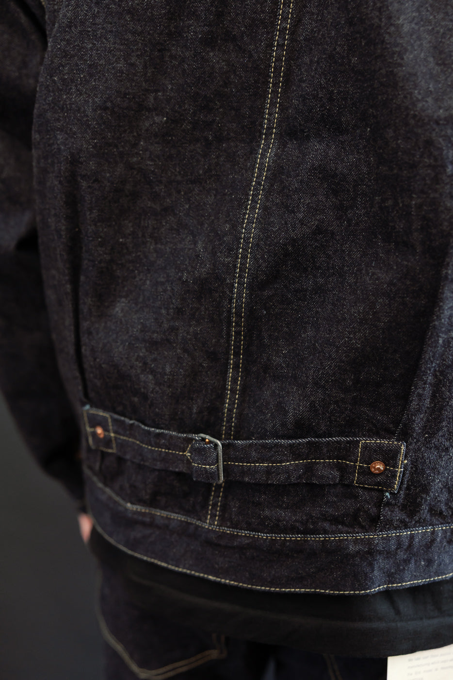 Close-up of back waistband on dark selvedge denim jacket, showing belt loops, copper rivets, and contrast stitching on male model.