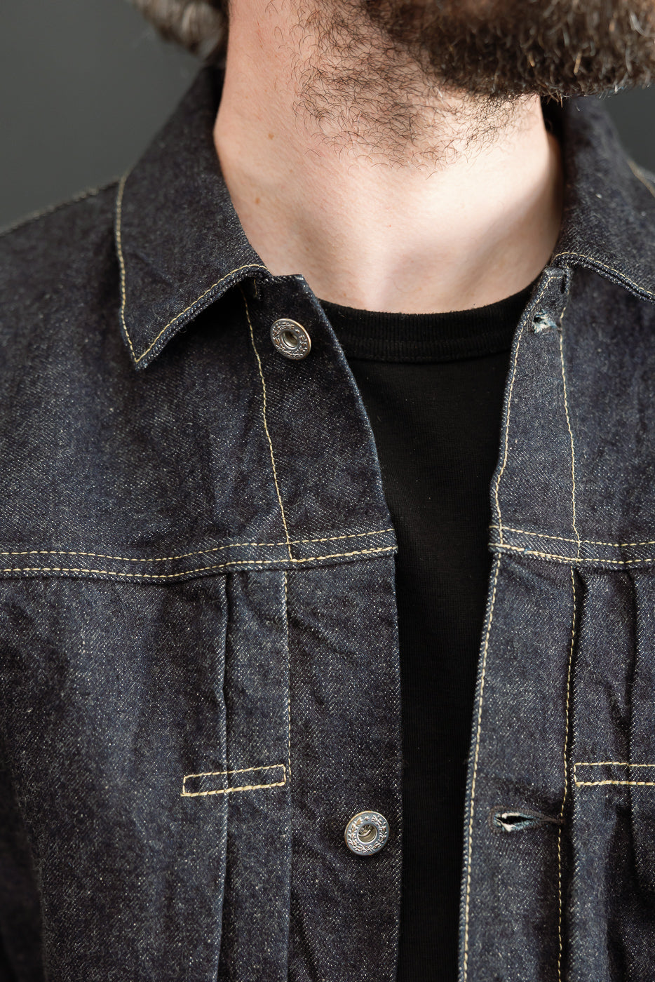 Close-up front view of male model wearing dark selvedge denim jacket with contrast stitching, button details, and chest pockets.