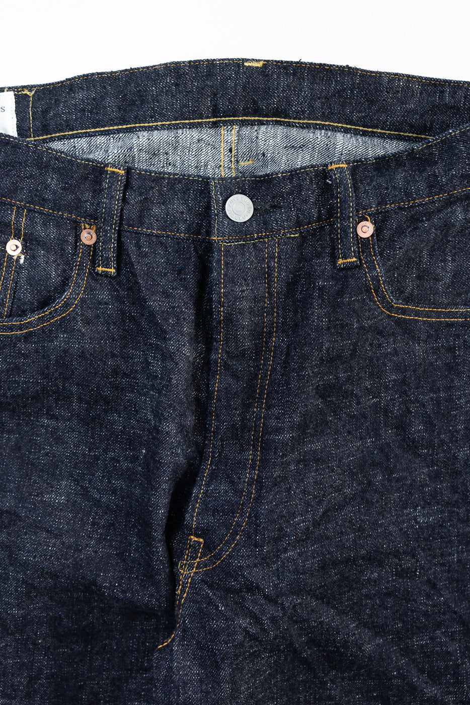 Close-up front view of raw selvedge denim jeans waist, showing button, rivets, and pocket stitching.
