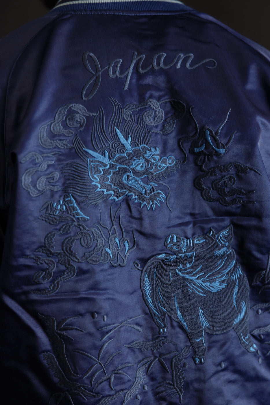 Close-up of the back of a male model’s indigo-dyed sukajan jacket, showing intricate dragon and boar embroidery and satin fabric texture. Model is 5'9