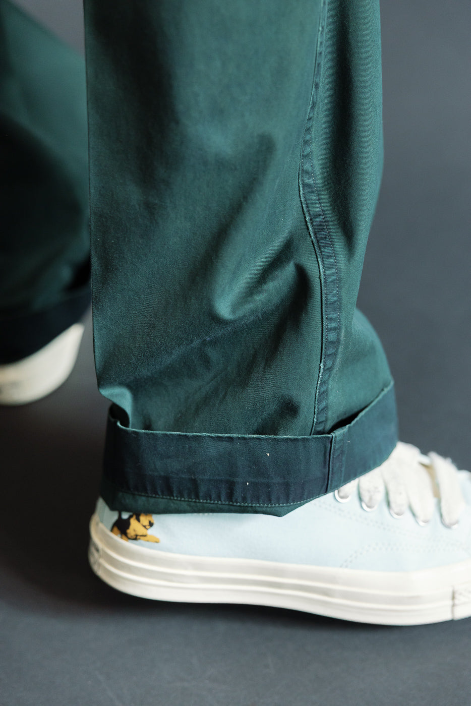 Close-up of male model’s cuffed dark green pants hem, showing fabric texture and stitching, paired with light blue sneakers