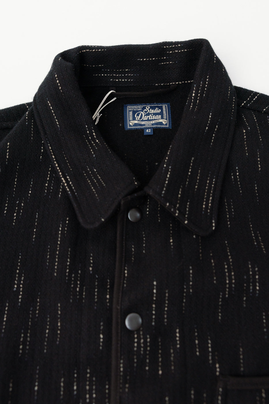 Close-up of Studio D'Artisan Kasuri-dyed black beach cloth coverall, front collar, snap buttons, textured fabric, visible tag.