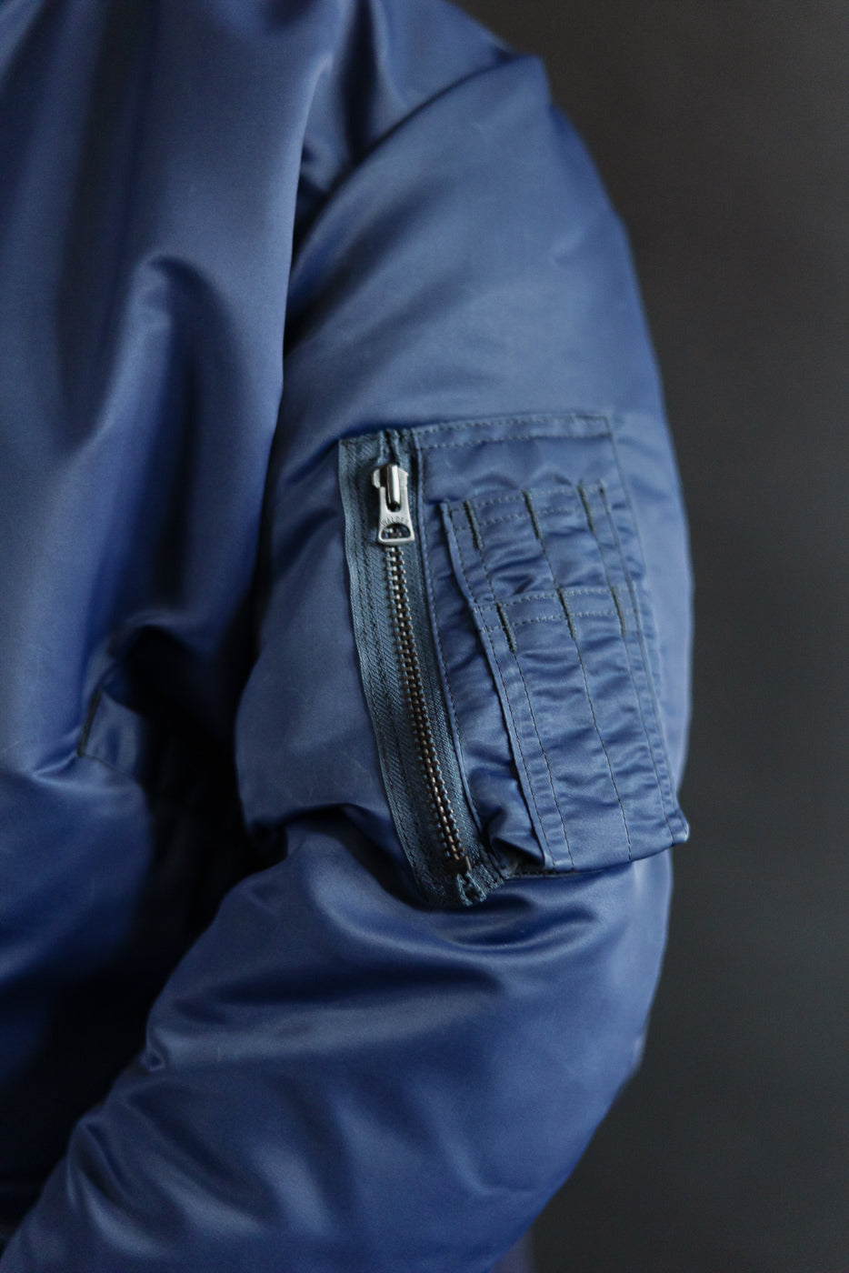 Close-up of indigo MA-1 flight jacket sleeve pocket zipper and stitching, men's outerwear detail