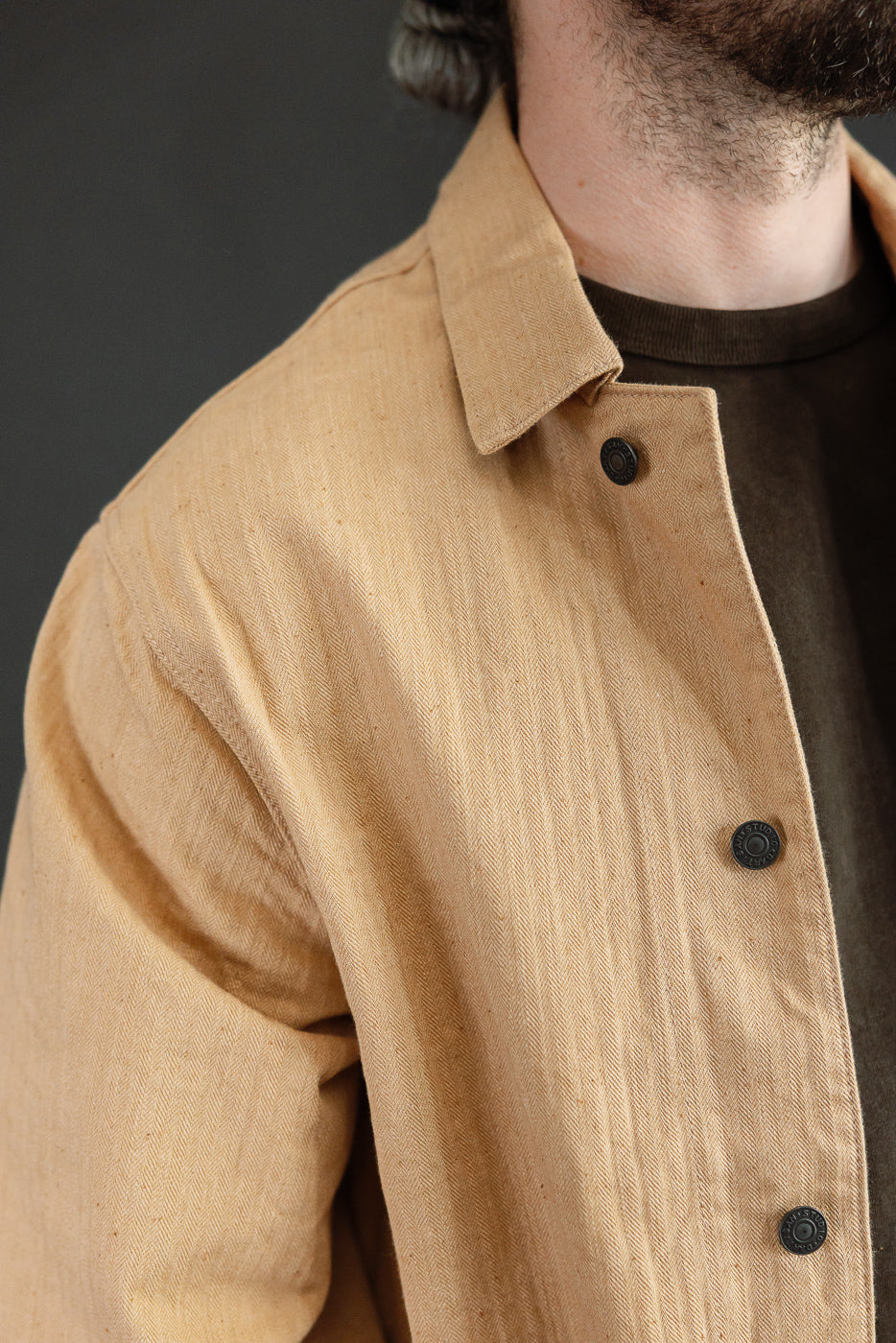 Close-up front view of tan herringbone chore jacket on male model, showing collar, black buttons, and fabric texture