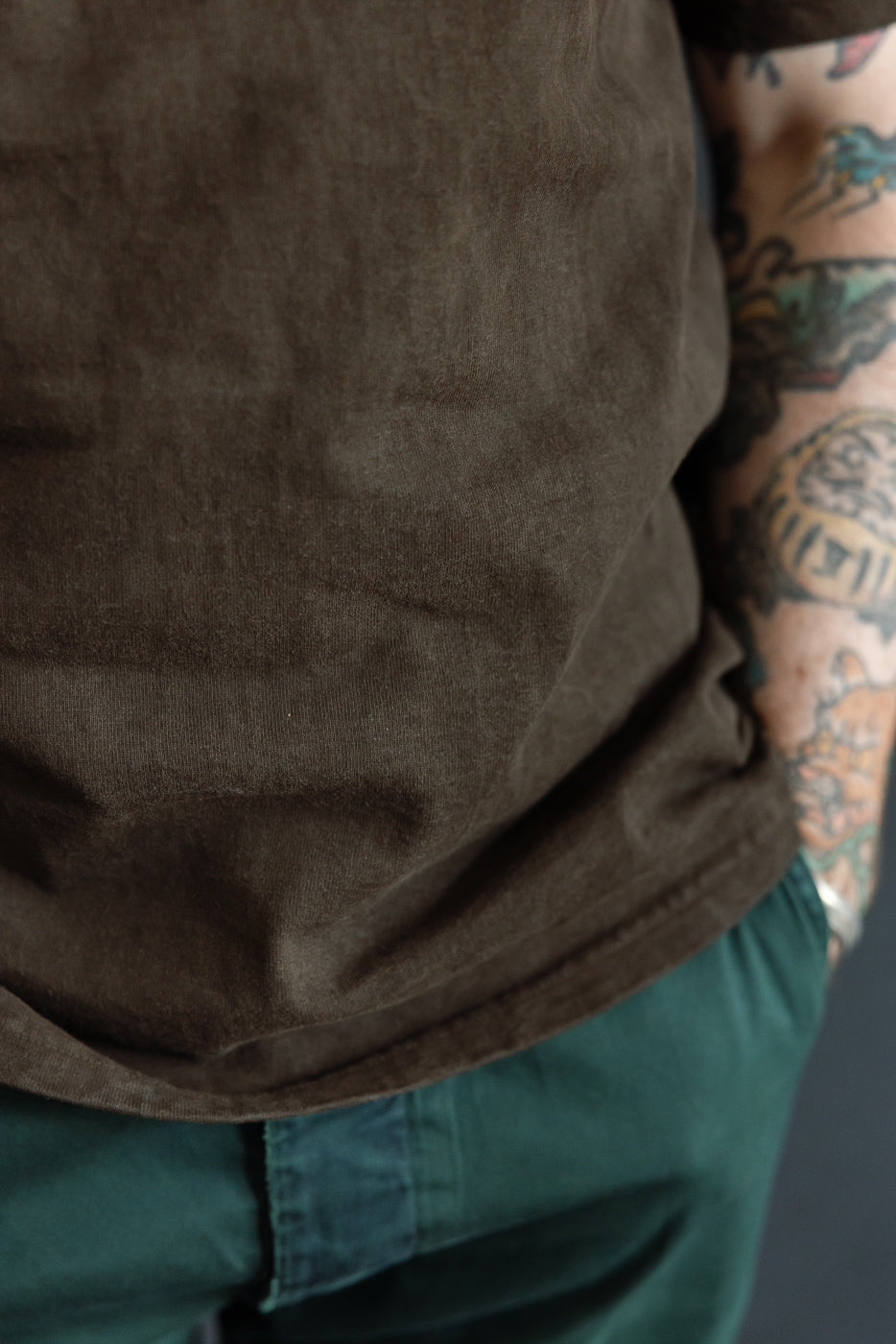 Close-up of olive t-shirt hem, worn by male model with tattoos, paired with green pants.
