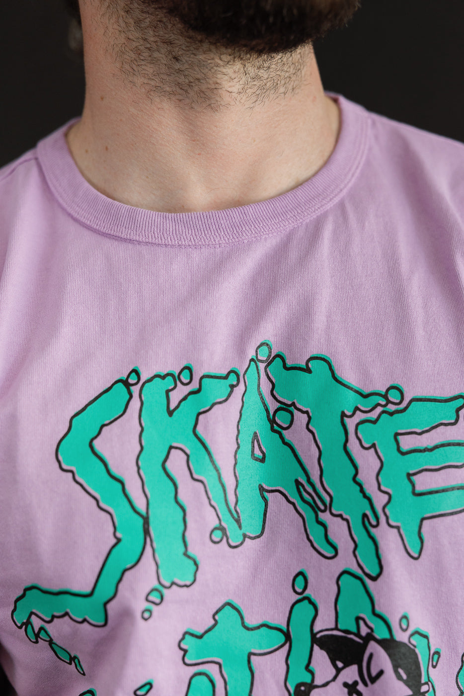Close-up front view of male model wearing lavender tee with teal 'SKATE' graphic, ribbed crew neck, athletic build