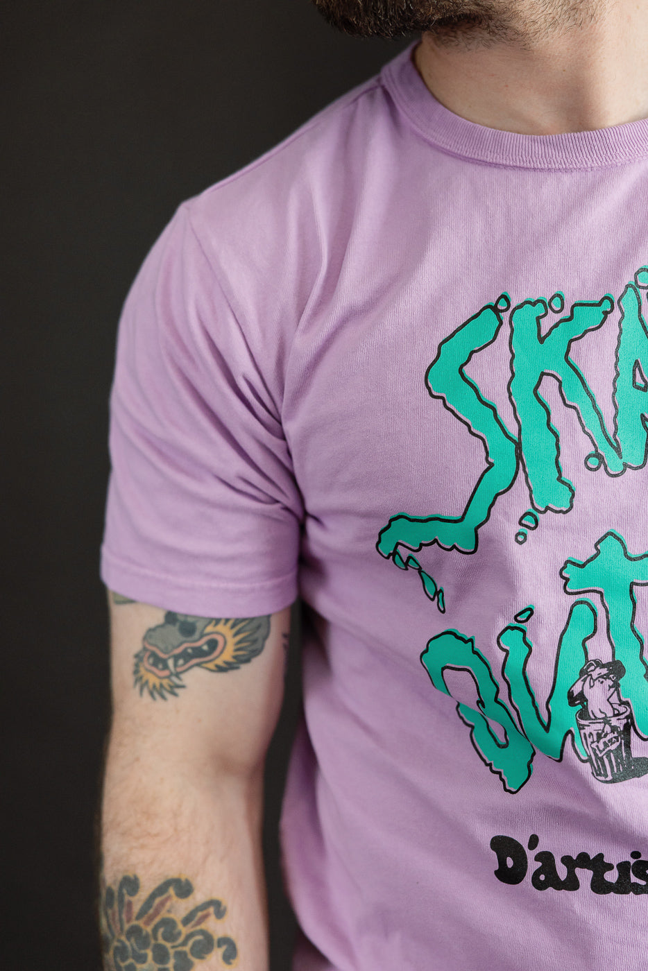 Close-up front view of male model wearing lavender tee with green graphic, showcasing sleeve and chest design.