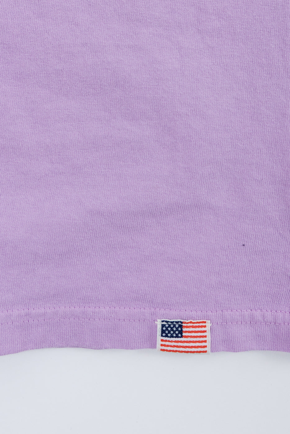 Close-up of lavender t-shirt hem with American flag tag detail, showcasing fabric texture and stitching.