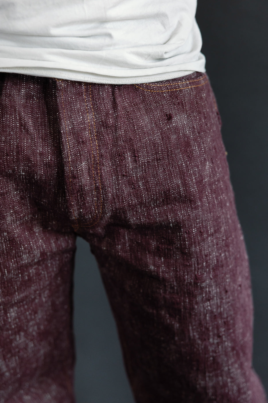 Close-up front view of maroon selvedge jeans, showcasing texture and orange stitching on male model.