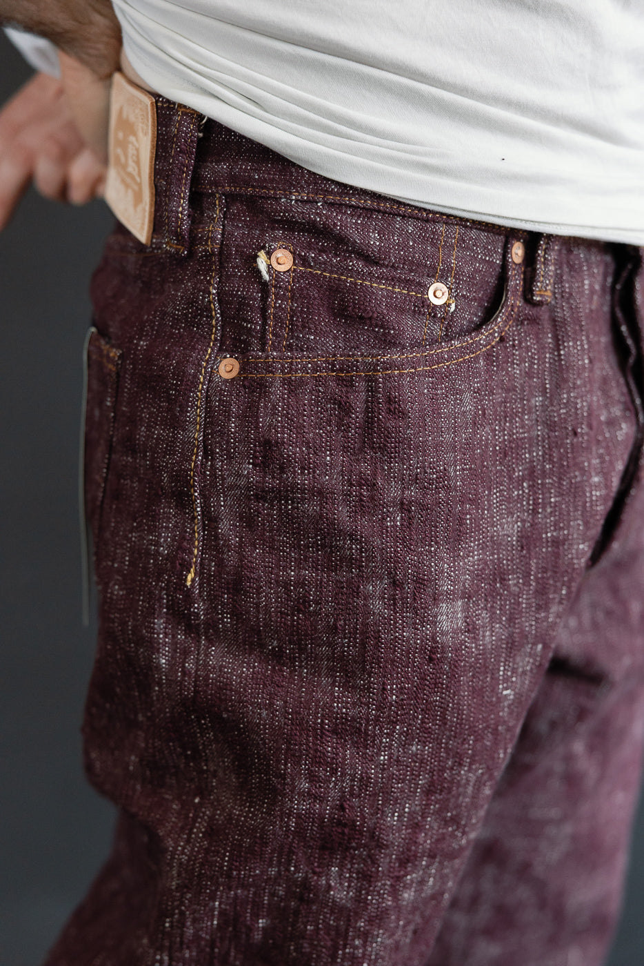Close-up front view of dark burgundy selvedge jeans with copper buttons and stitching, worn by male model.
