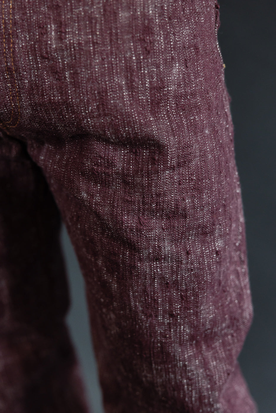 Close-up of maroon selvedge jeans leg, showcasing fabric texture and orange stitching detail.