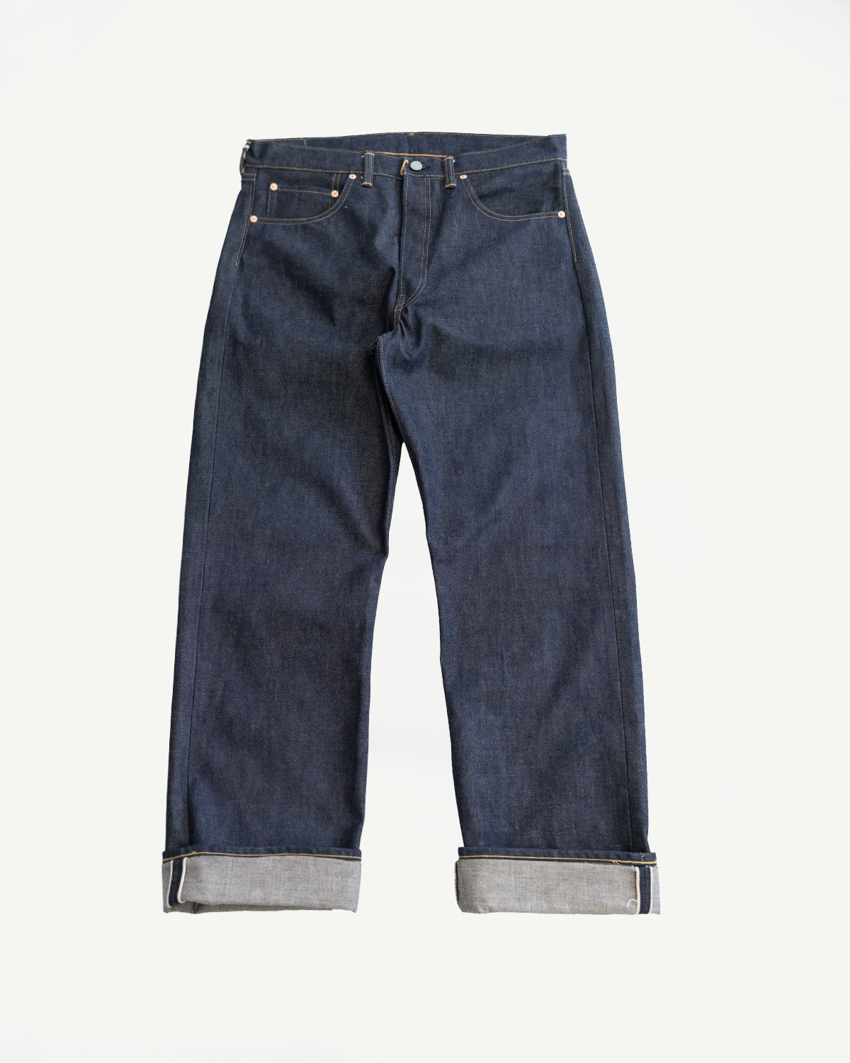 Flat lay of raw indigo selvedge denim jeans, front side, showcasing button fly and cuffed hem.