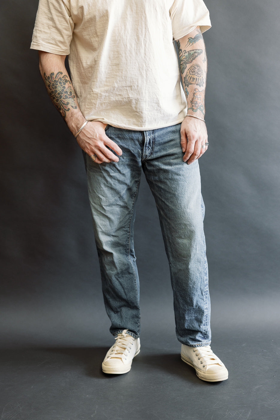 Person wearing Lot 2nd-Hand 1105 - 12oz Used Washed Indigo Denim - Tapered Fit, featuring vintage-style mid-wash jeans with a relaxed waist, slightly higher rise, and gentle taper, styled with white canvas sneakers.