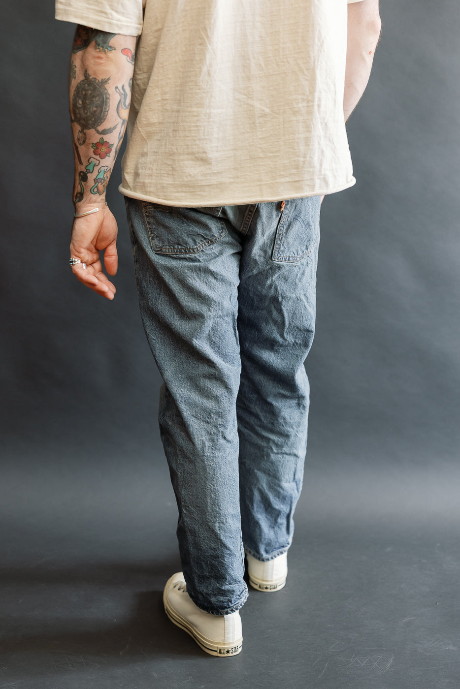 Back view of the Lot 2nd-Hand 1105 - 12oz Used Washed Indigo Denim - Tapered Fit jeans, featuring relaxed-fit blue denim with a tapered silhouette, paired with white canvas sneakers and off-white T-shirt.