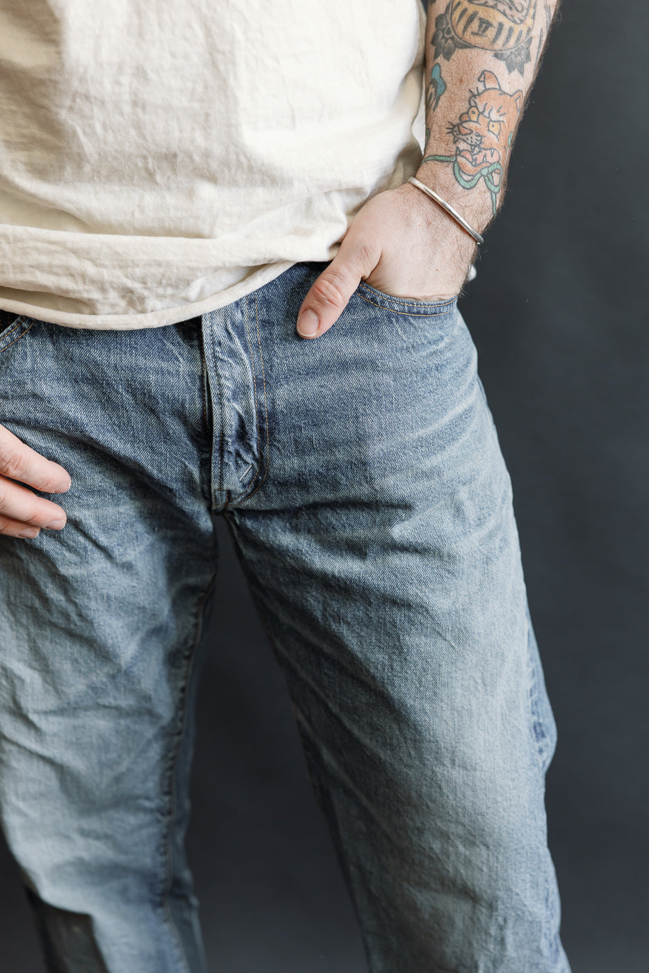 Close-up of Lot 2nd-Hand 1105 - 12oz Used Washed Indigo Denim - Tapered Fit, featuring slightly wrinkled off-white T-shirt tucked into faded blue jeans, showcasing colorful tattooed forearm with silver cuff bracelet.