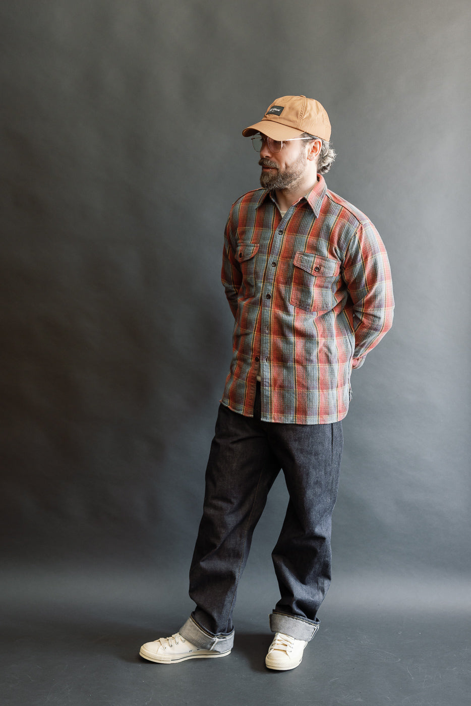 Man wearing Lot 3022 - Flannel Shirt w/ Chinstrap G Pattern - Red, Navy, styled with dark cuffed denim and off-white sneakers, capturing heritage workwear aesthetics. Hands clasped, he gazes slightly aside.