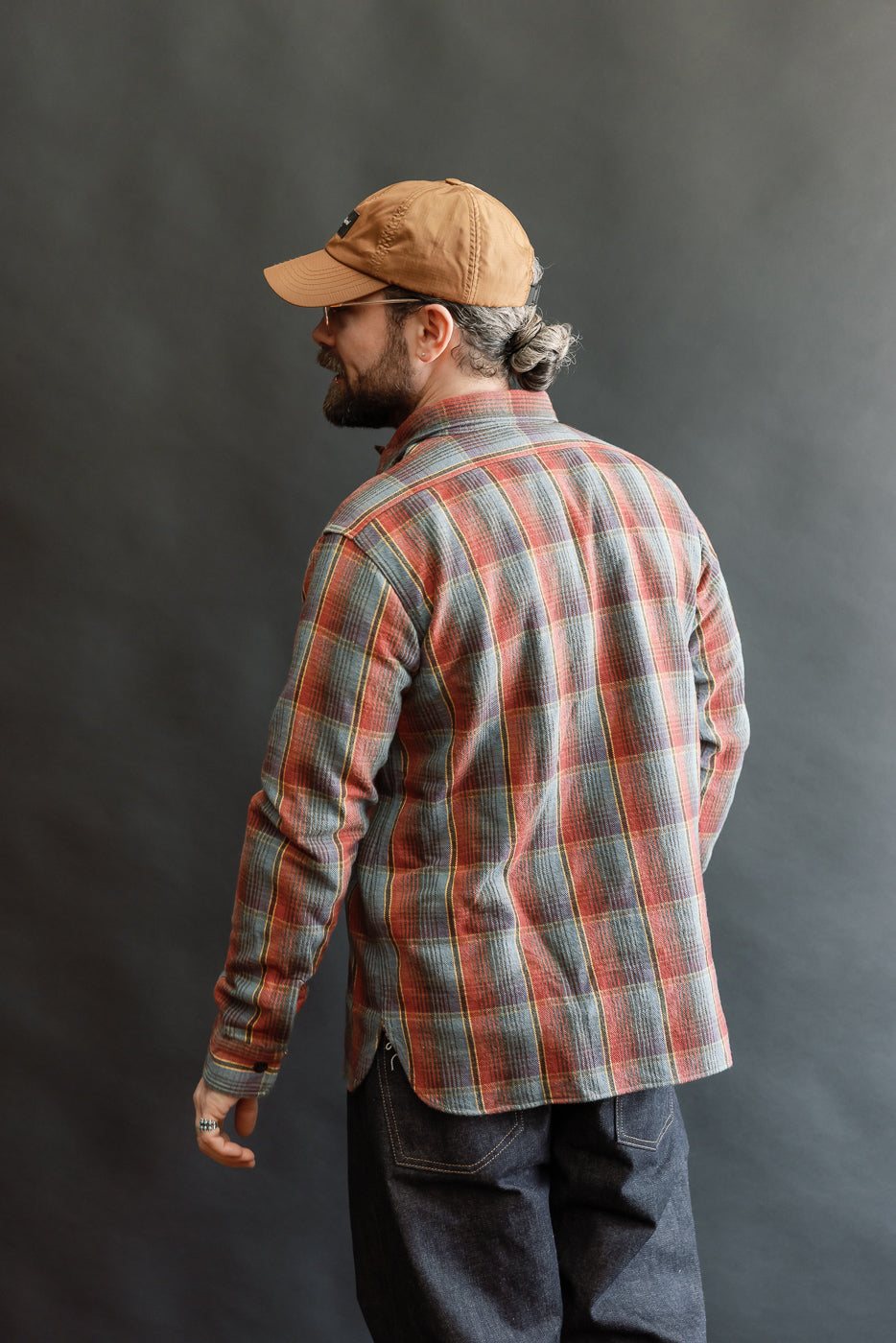 Person facing away, wearing The Warehouse & Co. Lot 3022 Flannel Shirt w/ Chinstrap G Pattern in red and navy plaid, showcasing its vintage-inspired design, paired with dark denim jeans and a brown baseball cap.