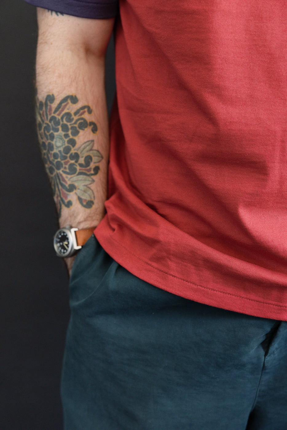 Close-up, side view of a male model's arm with a floral tattoo wearing a red t-shirt and dark trousers.