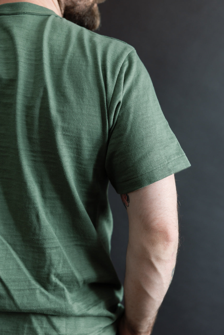 Close-up back view of green slubby cotton t-shirt on male model, 5'9, 165lbs, athletic build, showing sleeve and shoulder texture.
