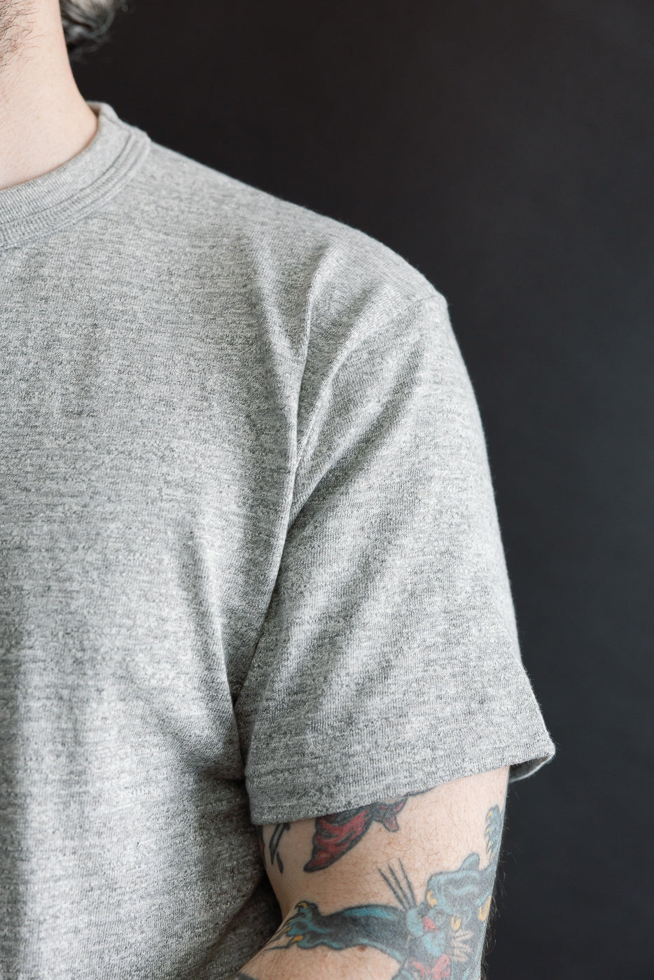 Close-up of male model's right sleeve and shoulder of heather grey slubby cotton T-shirt, athletic build, showing fabric texture and classic crew neck.