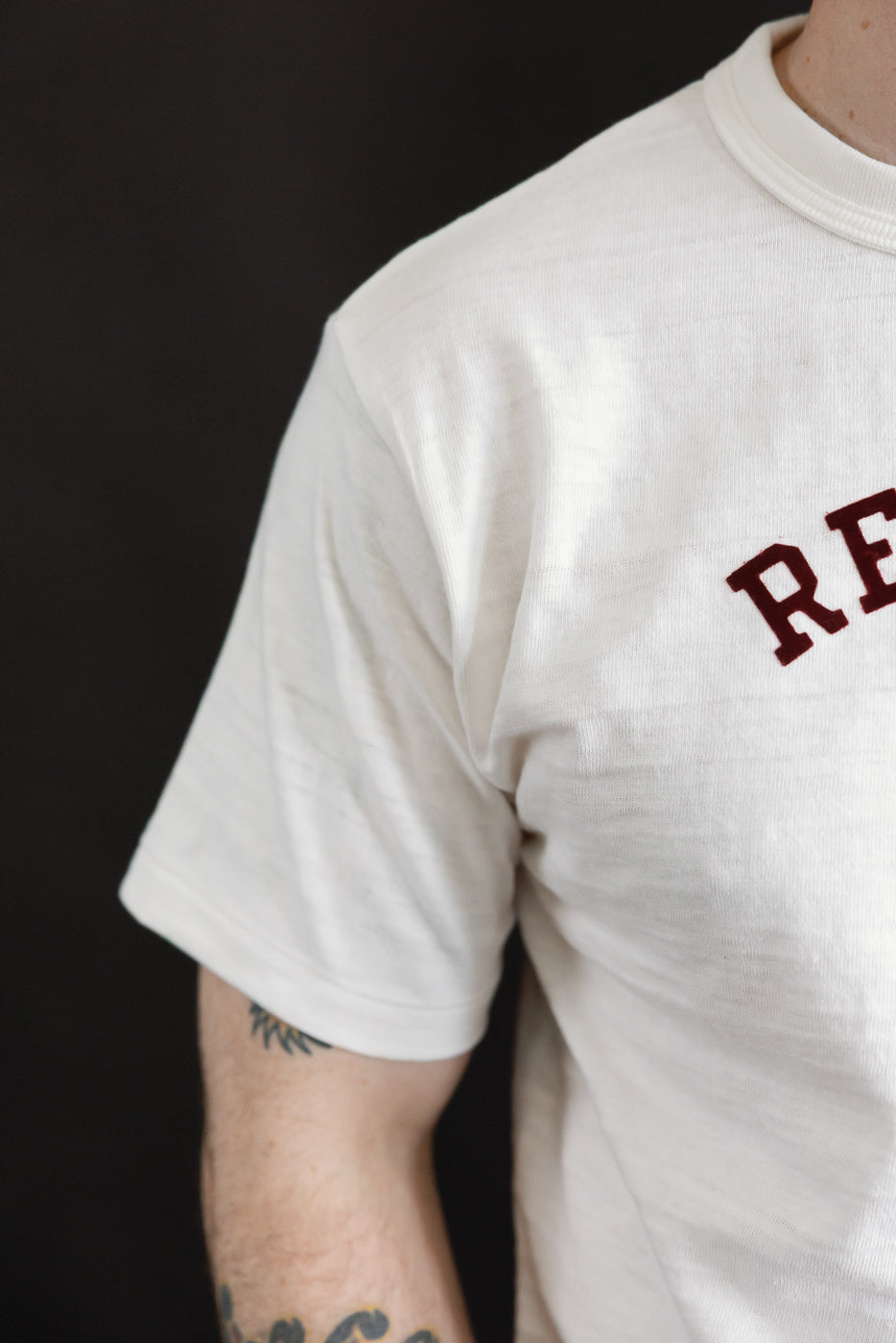 Close-up of male model’s right front shoulder wearing Warehouse & Co slubby cotton off-white T-shirt, showing texture, ribbed collar, and partial 'RE' red lettering. Model is 5'9
