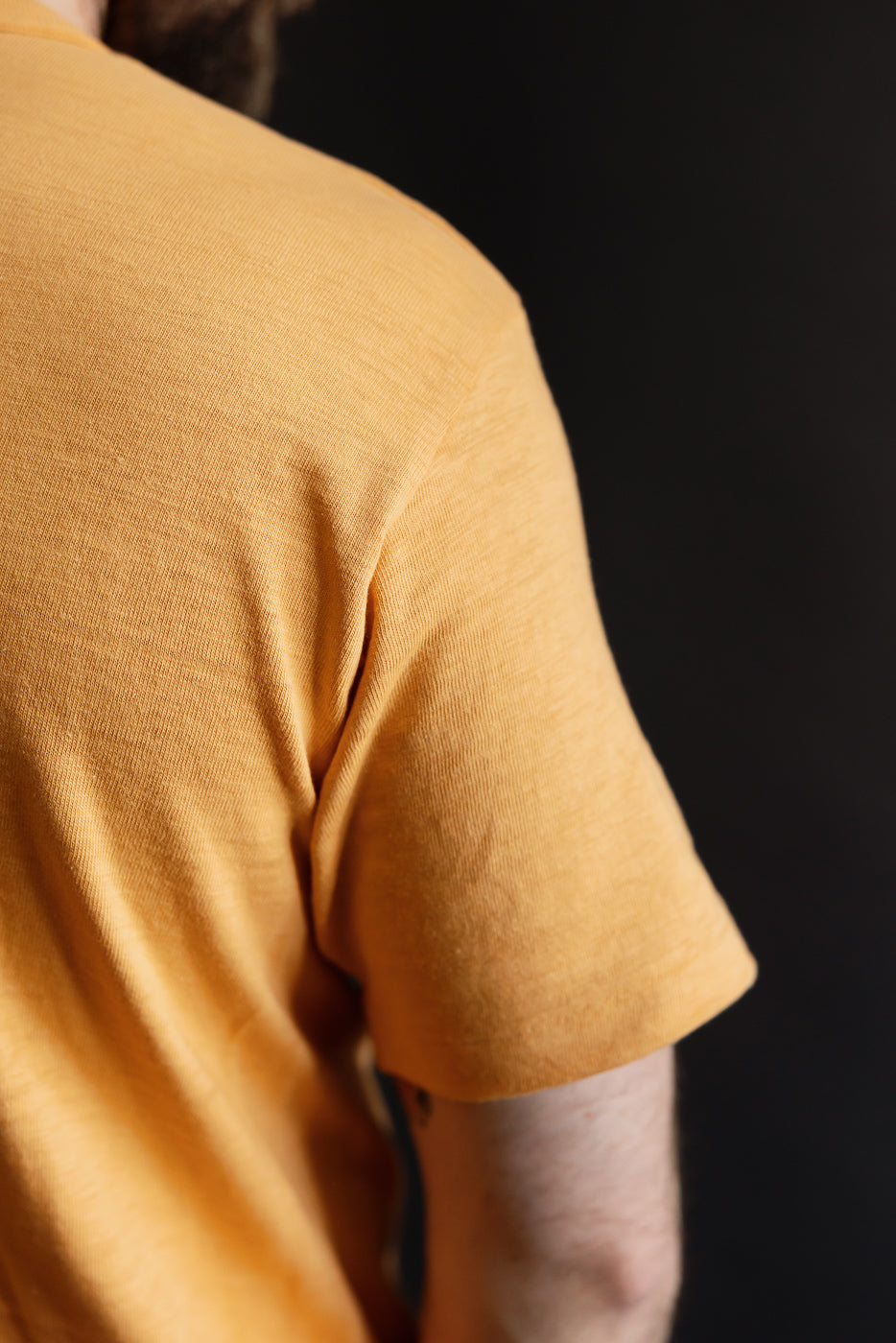 Close-up of male model's upper back and sleeve in orange slubby cotton t-shirt, left rear view
