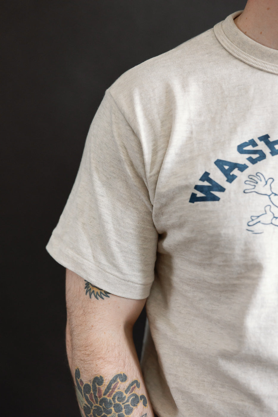 Close-up of male model's left side wearing Warehouse & Co. slubby oatmeal cotton T-shirt, front graphic, short sleeve, crewneck; model is 5'9