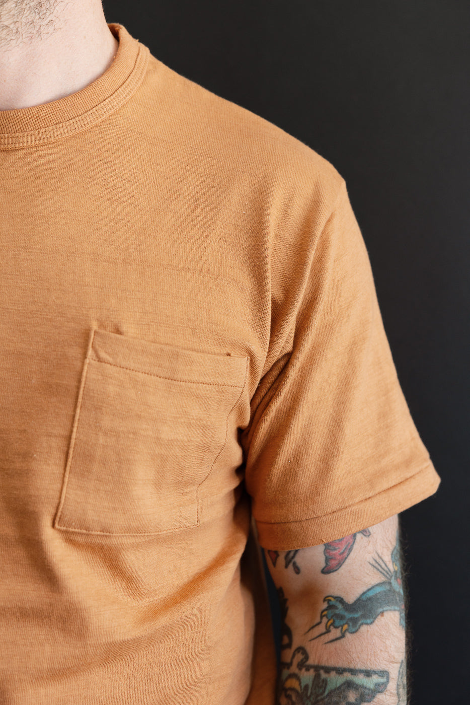 Close-up, front view of a tan pocket t-shirt showing the chest pocket and sleeve detail.