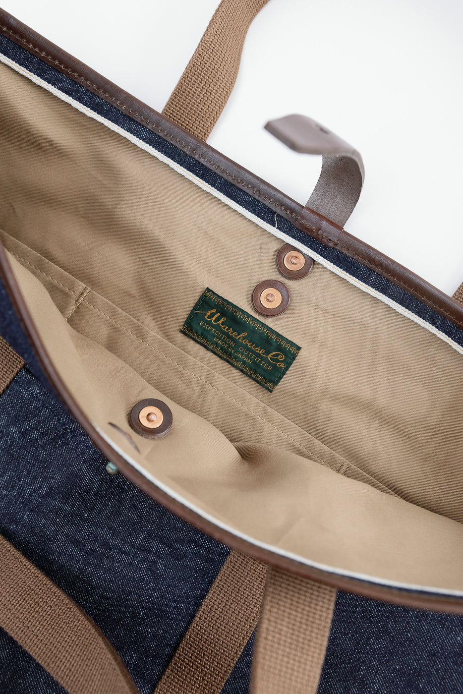 Close-up of interior top view of denim tote bag showing snap buttons, branded tag, and beige liner.