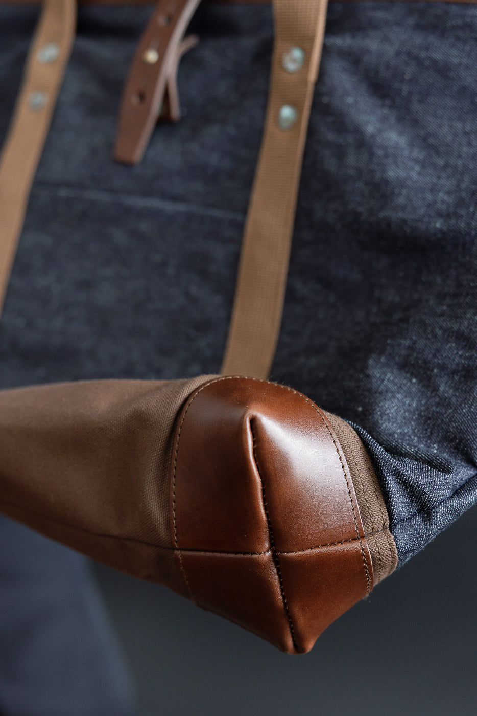 Close-up of denim tote bag bottom corner, showing brown leather reinforced base and riveted canvas straps.