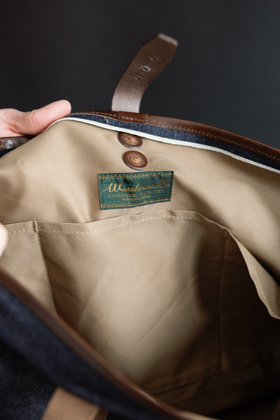 Close-up of denim tote bag interior, showing inner pocket, buttons, and brand tag detail.