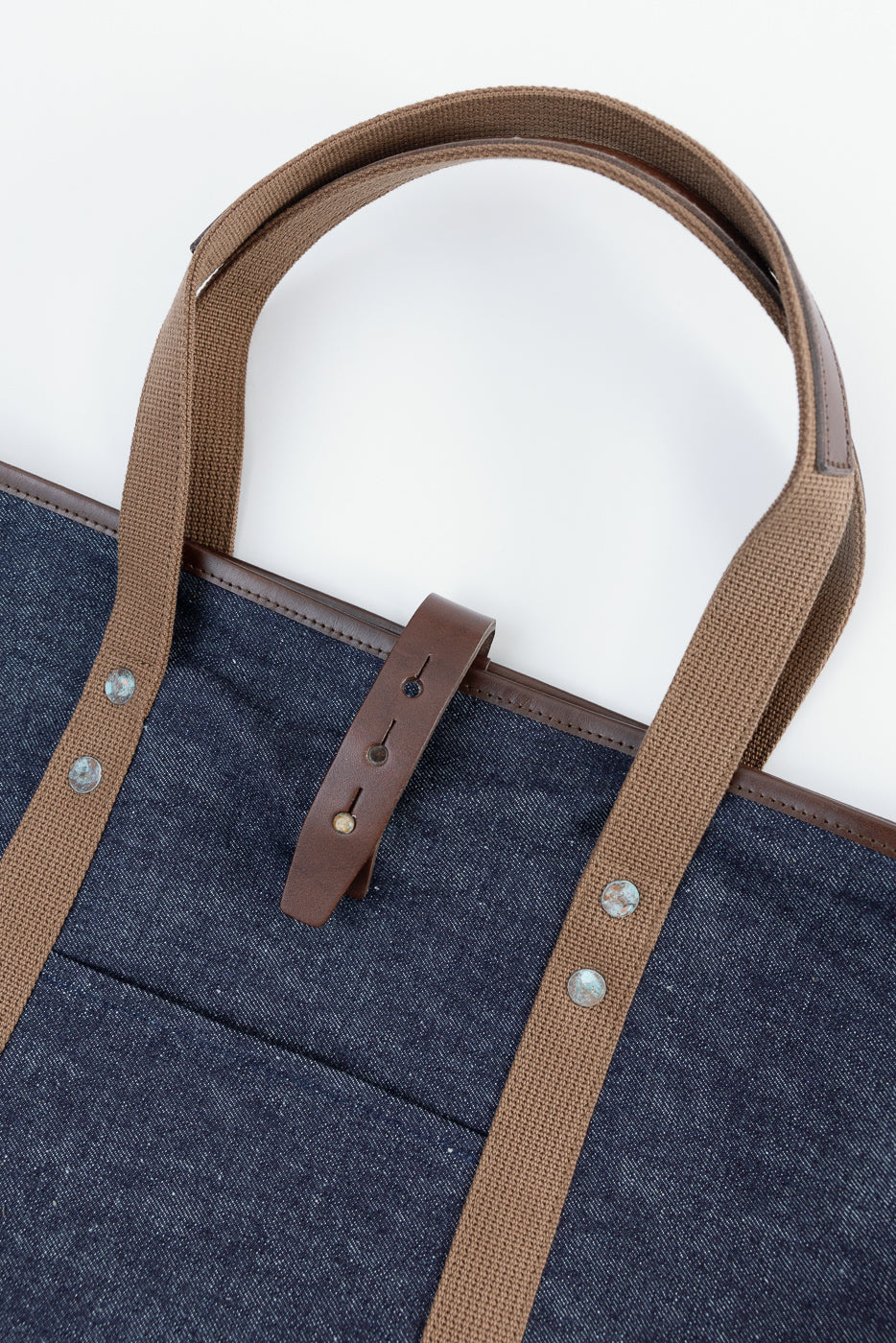 Close-up of denim tote bag front, showing brown canvas straps, leather tab, and rivet details.
