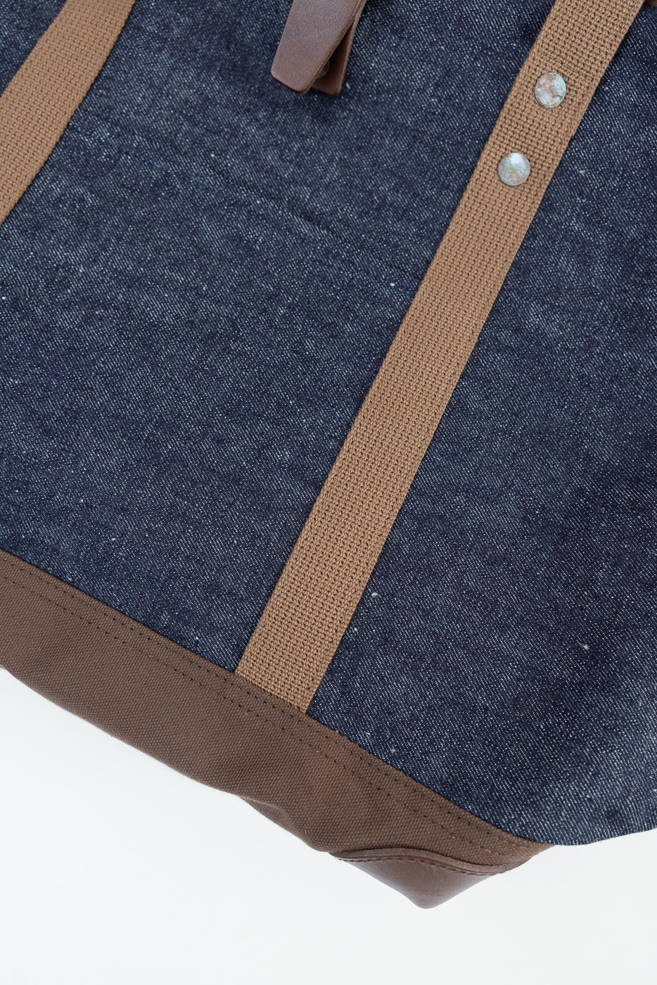 Close-up of front lower panel of indigo denim tote bag with brown straps and rivet details.