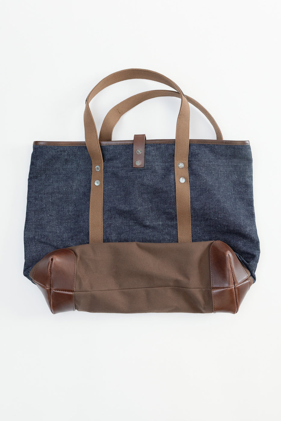 Flat lay, front view of denim and canvas tote bag with brown leather accents and straps.