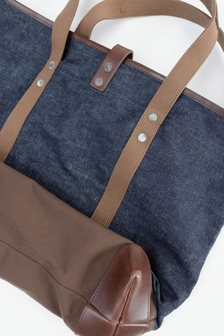 Close-up of denim tote bag front, showing brown canvas straps, leather trim, and rivet details.