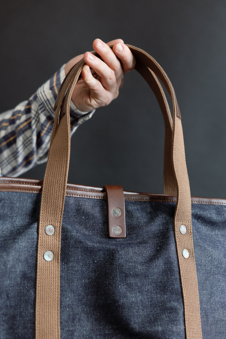 Close-up of front side of indigo denim tote bag with brown straps, male hand holding handle, showing rivet and leather tab details.