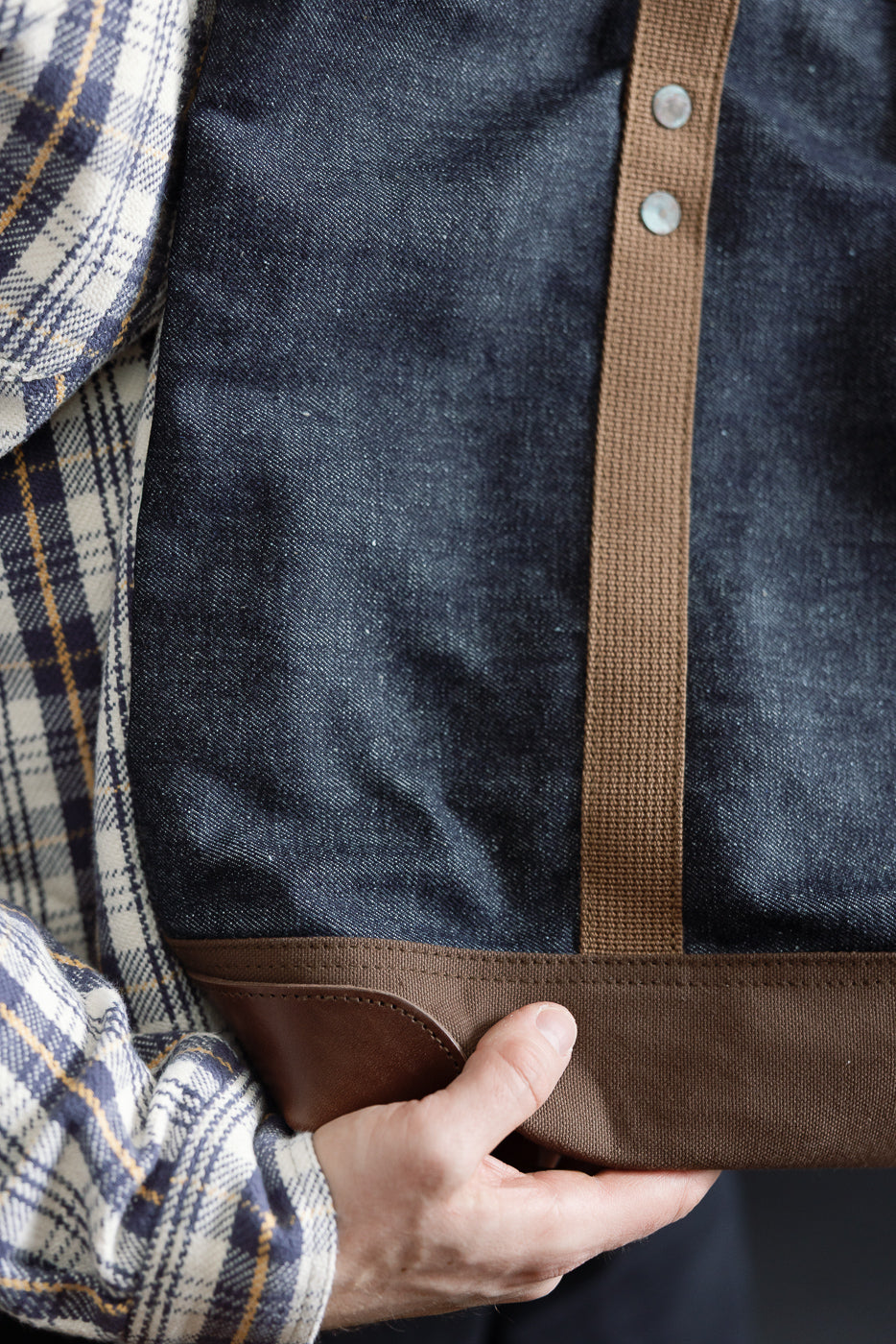 Close-up front view of indigo denim tote bag with brown canvas trim and rivet details, held by male model, 5'9